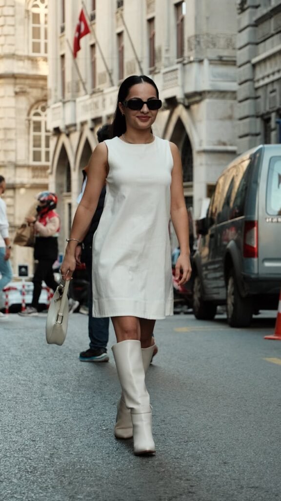Elegant woman in white dress and boots walking confidently in city street.